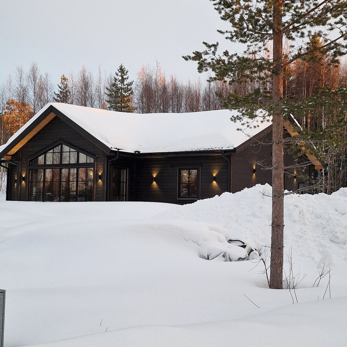 A modern cabin is set against a snowy landscape, featuring a dark exterior with large windows. The roof is blanketed in snow, while soft outdoor lighting enhances the building's architectural lines. Pine trees frame the view, completing the serene winter scene.
