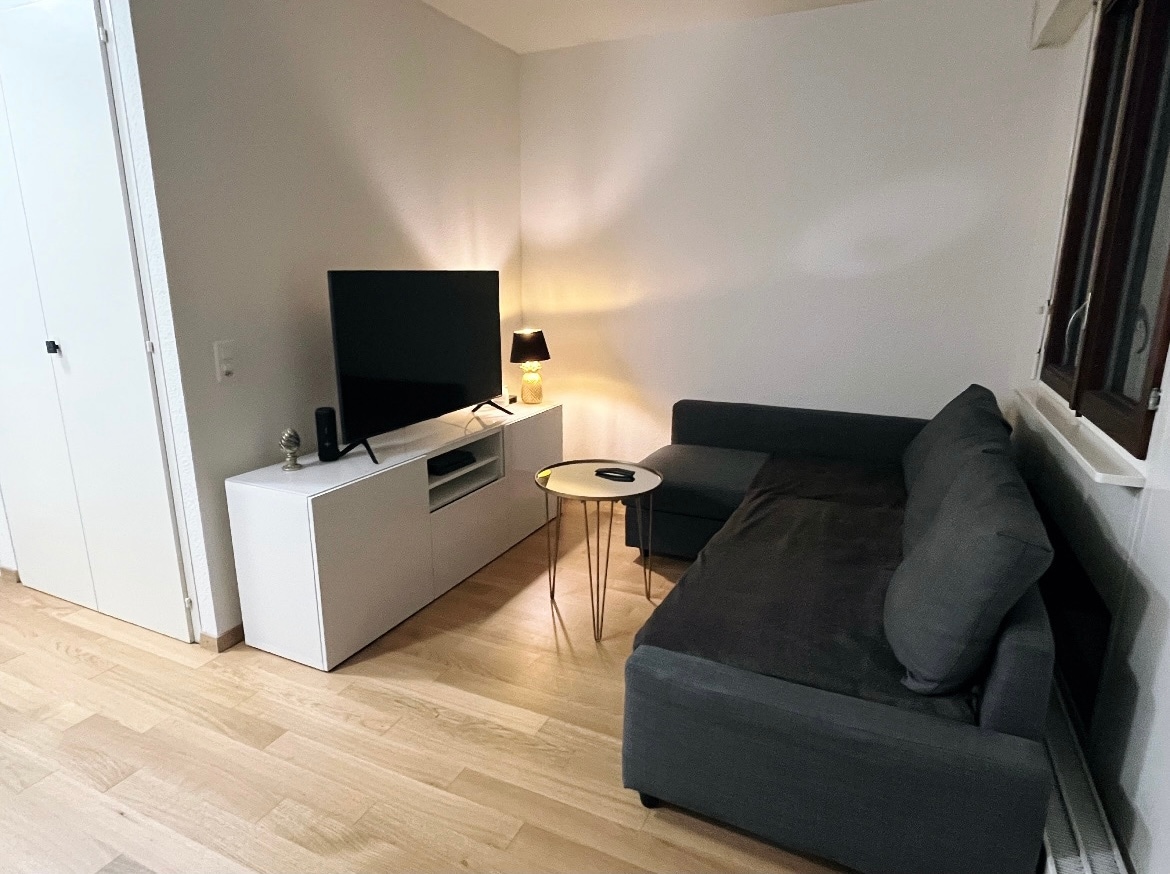 A cozy living area features a dark gray sofa with a chaise section and a small round glass coffee table. A flat-screen TV is mounted on a white media console. Soft lighting is provided by a lamp on the console, creating a relaxing ambiance.