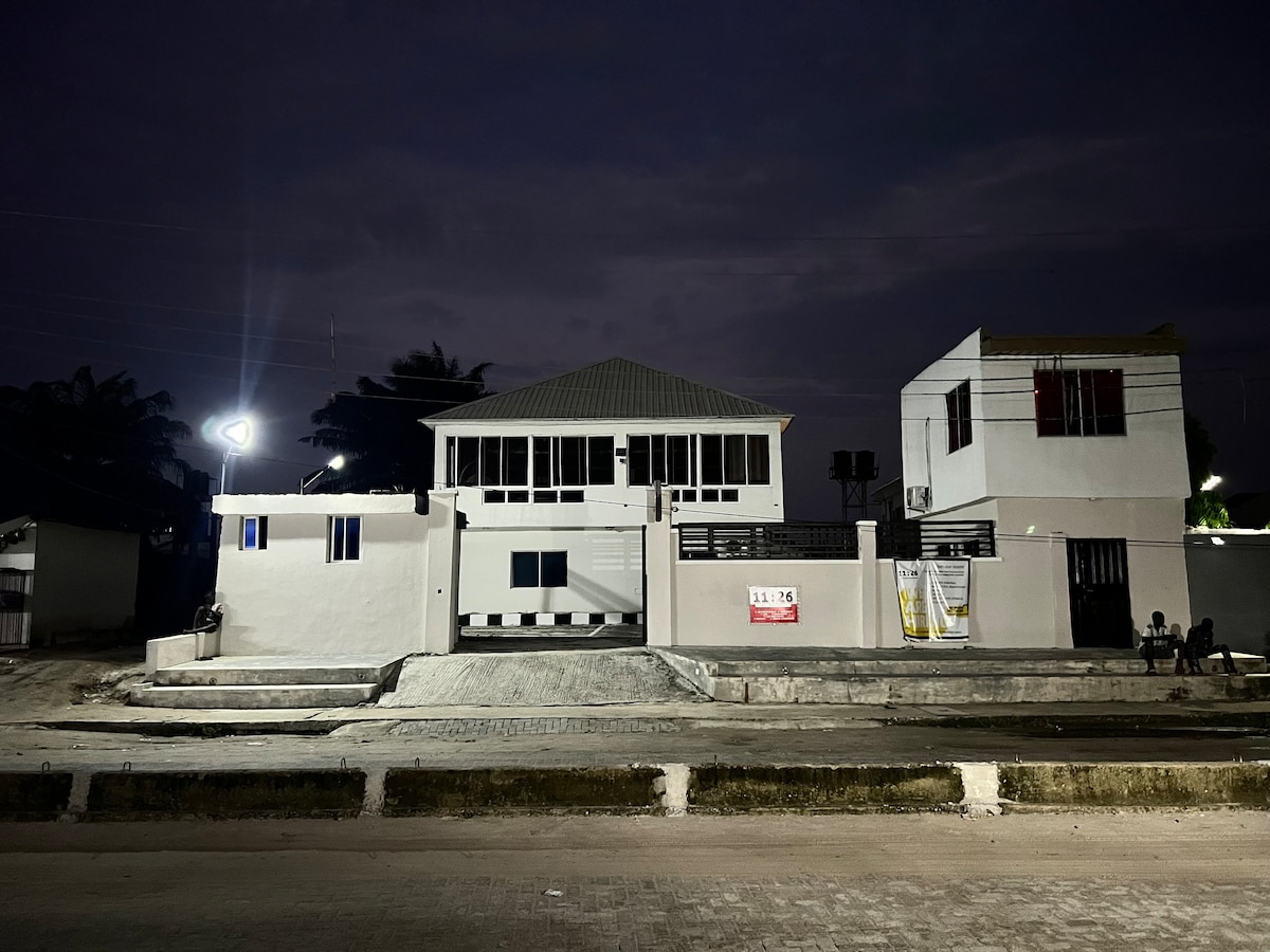 The modern building is illuminated against a dark sky, showcasing a combination of white walls and large windows. A covered porch extends from the main entrance, providing a spacious area. The surrounding environment features a pathway and streetlight, contributing to a well-defined entryway.