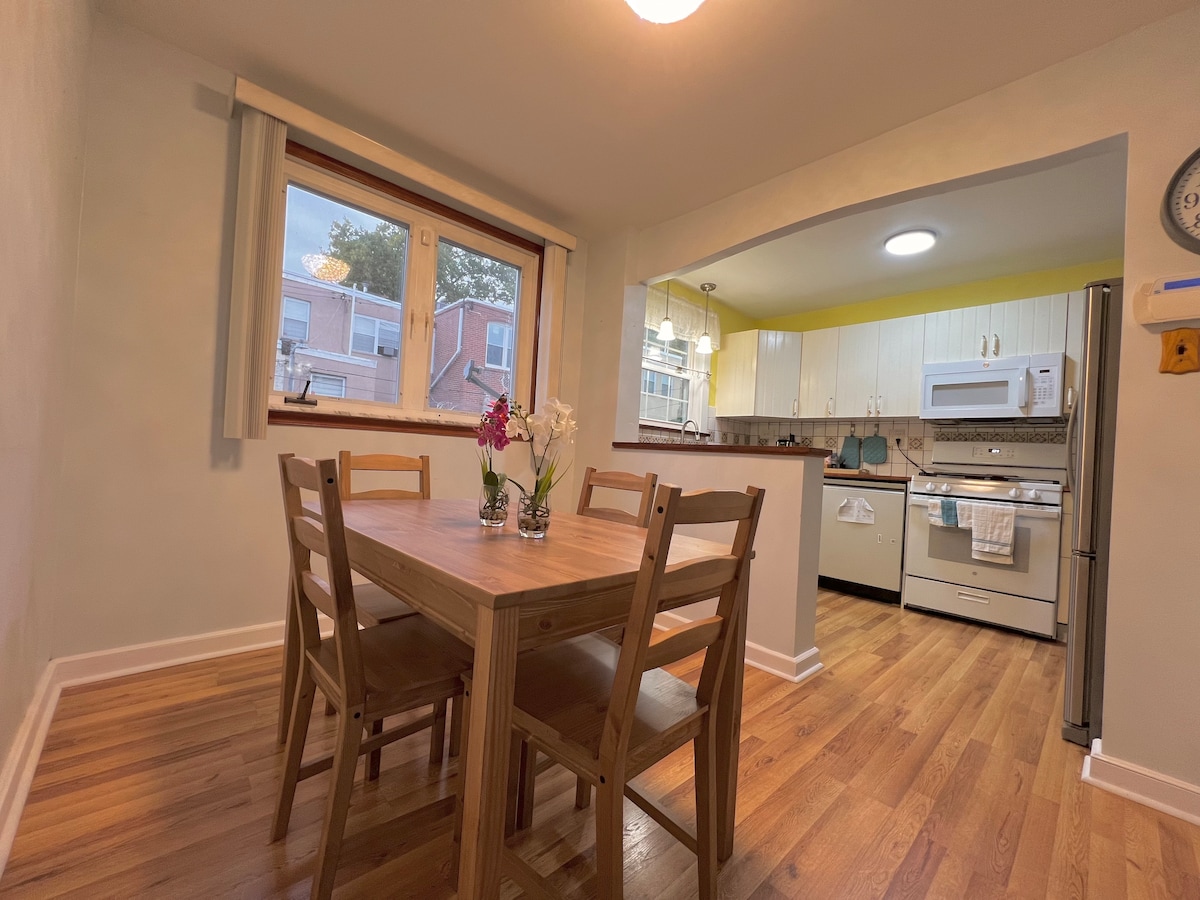 A dining area features a wooden table surrounded by four chairs. Natural light streams through a large window, illuminating a kitchen equipped with white cabinetry and modern appliances, including a stove and microwave. Decorative flowers are placed in two vases on the table.