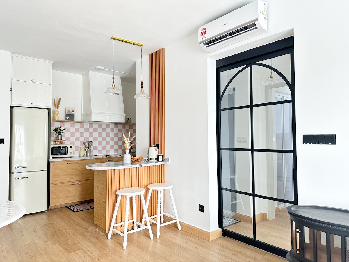 A modern kitchen area features light wood cabinetry with a kitchen island complemented by three white stools. Soft pink tiles add a touch of color. An air conditioning unit is mounted above, and large glass doors provide a view of the interior space.