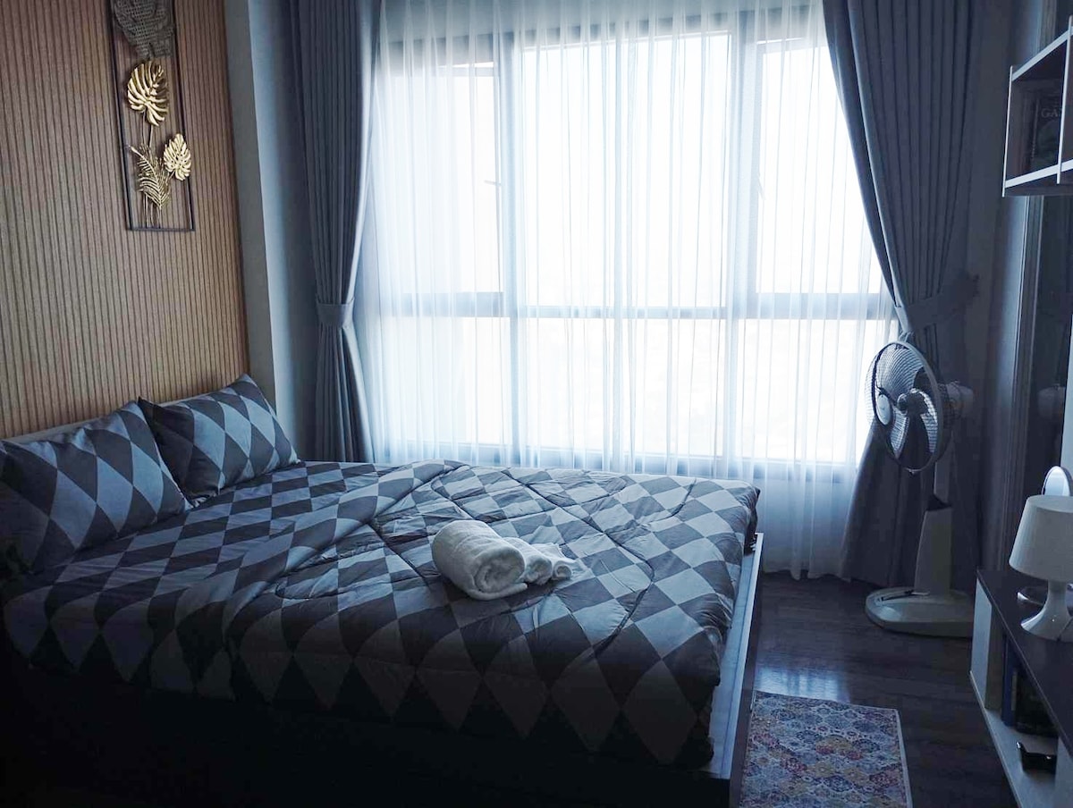 A cozy bedroom features a spacious bed dressed in a patterned quilt and multiple decorative pillows. Natural light floods the room through large windows covered with sheer curtains. A standing fan and a small bedside lamp are positioned nearby, enhancing the room's functionality.