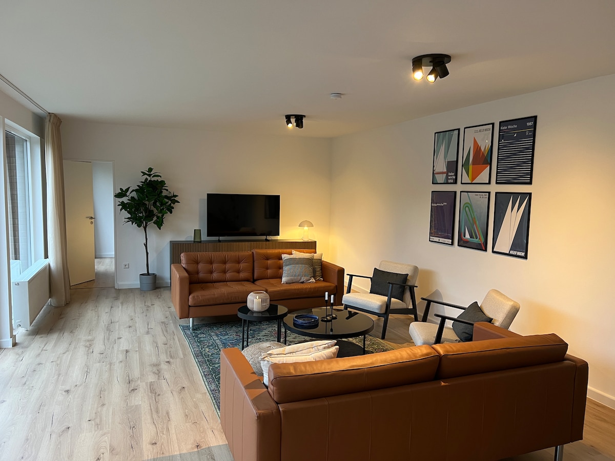 The spacious living area features a combination of comfortable sofas and armchairs arranged around a low coffee table. Artwork adorns the walls, and a flat-screen TV is mounted above a stylish console. Natural light fills the room, highlighting the modern decor.