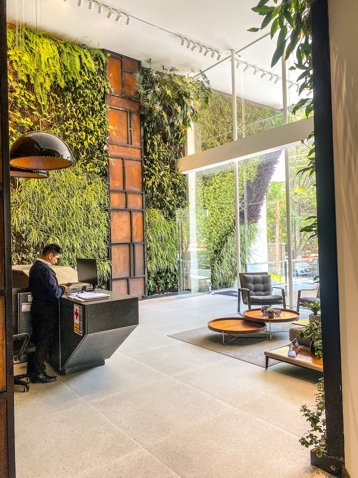 A welcoming reception area is presented with a blend of natural greenery and modern design. A large vertical garden features lush plants against a textured wall. A sleek front desk is situated to the left, with comfortable seating arranged in a cozy corner.