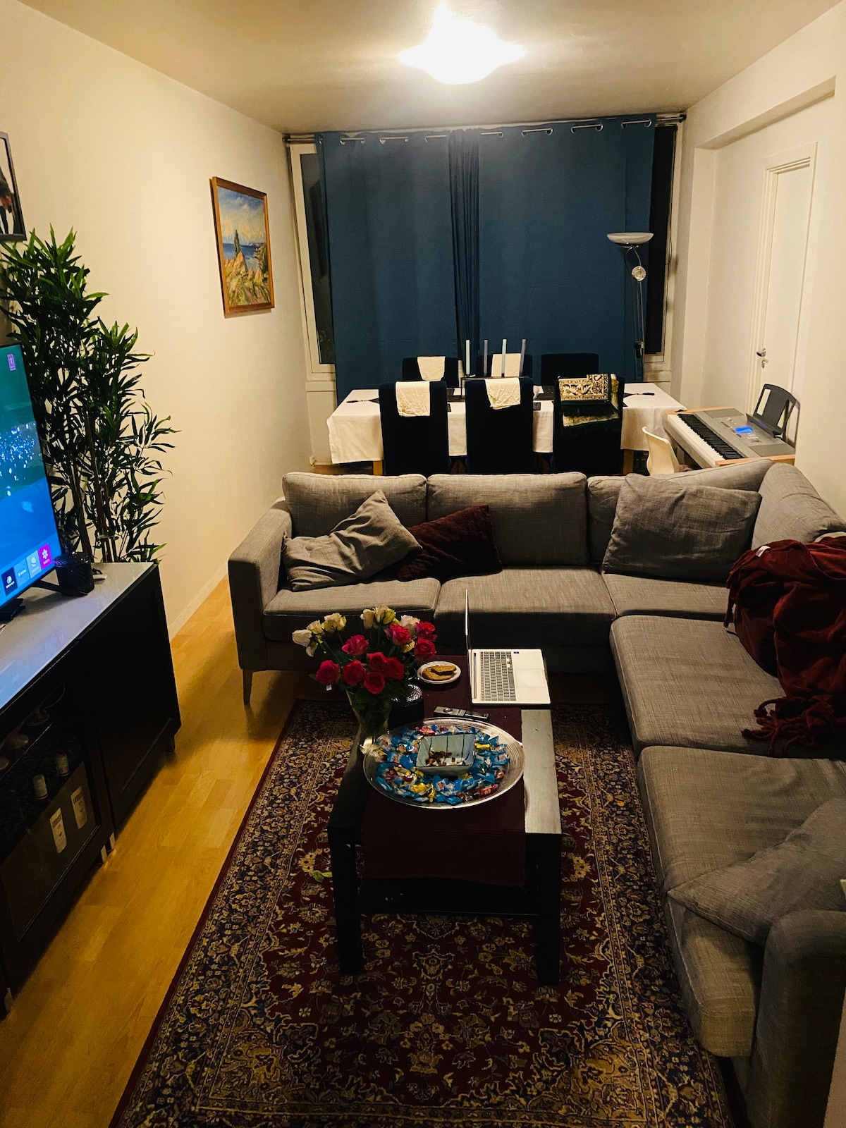 A section of the living area is furnished with a gray sectional sofa and a small table, adorned with a tray and a floral arrangement. A television is mounted on a dark cabinet, while a dining area with dark chairs and a piano is visible in the background. Soft lighting is provided by a ceiling fixture.