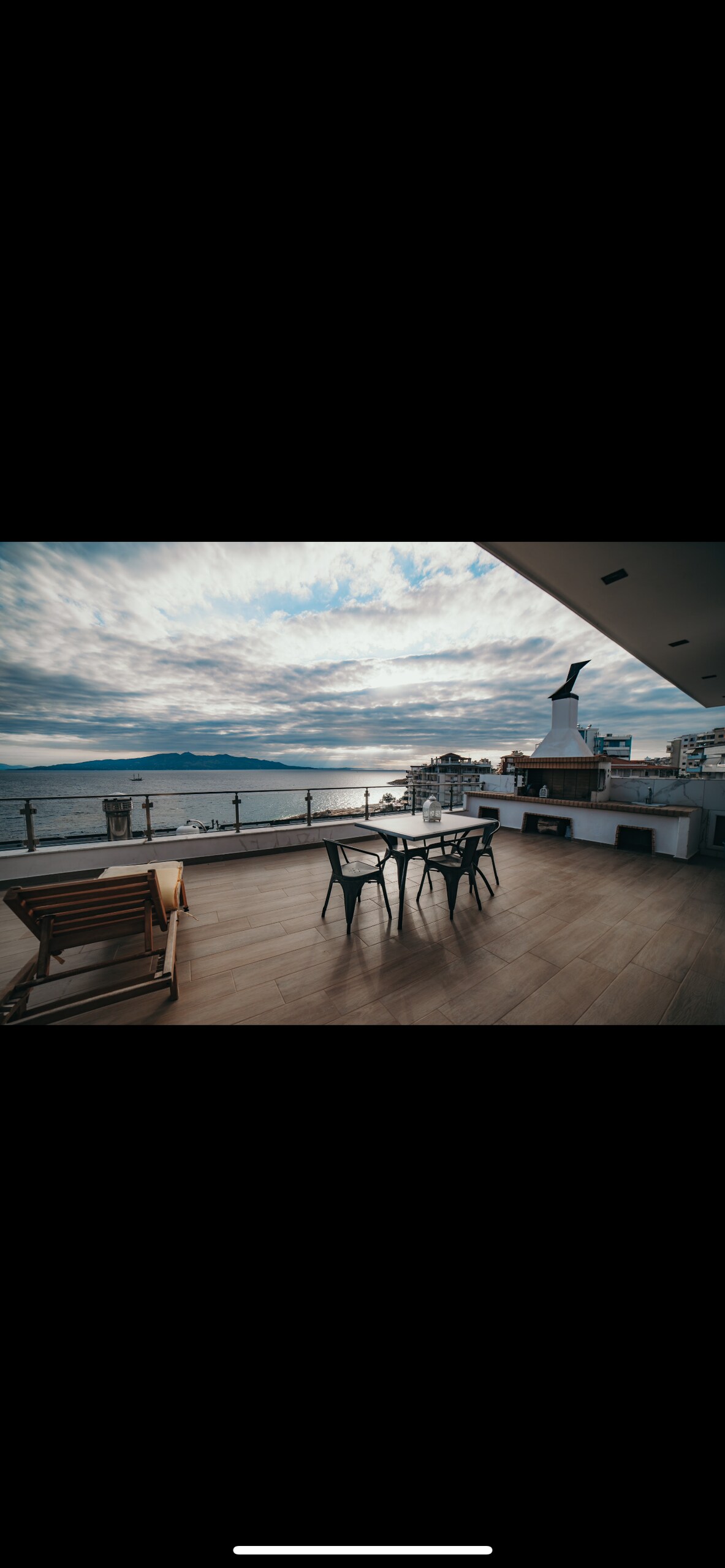 An outdoor terrace is featured with a table and chairs, set against a backdrop of a scenic ocean view under a cloudy sky. A lounge chair is positioned nearby, and a fireplace is integrated into the structure, adding a functional element to the space.