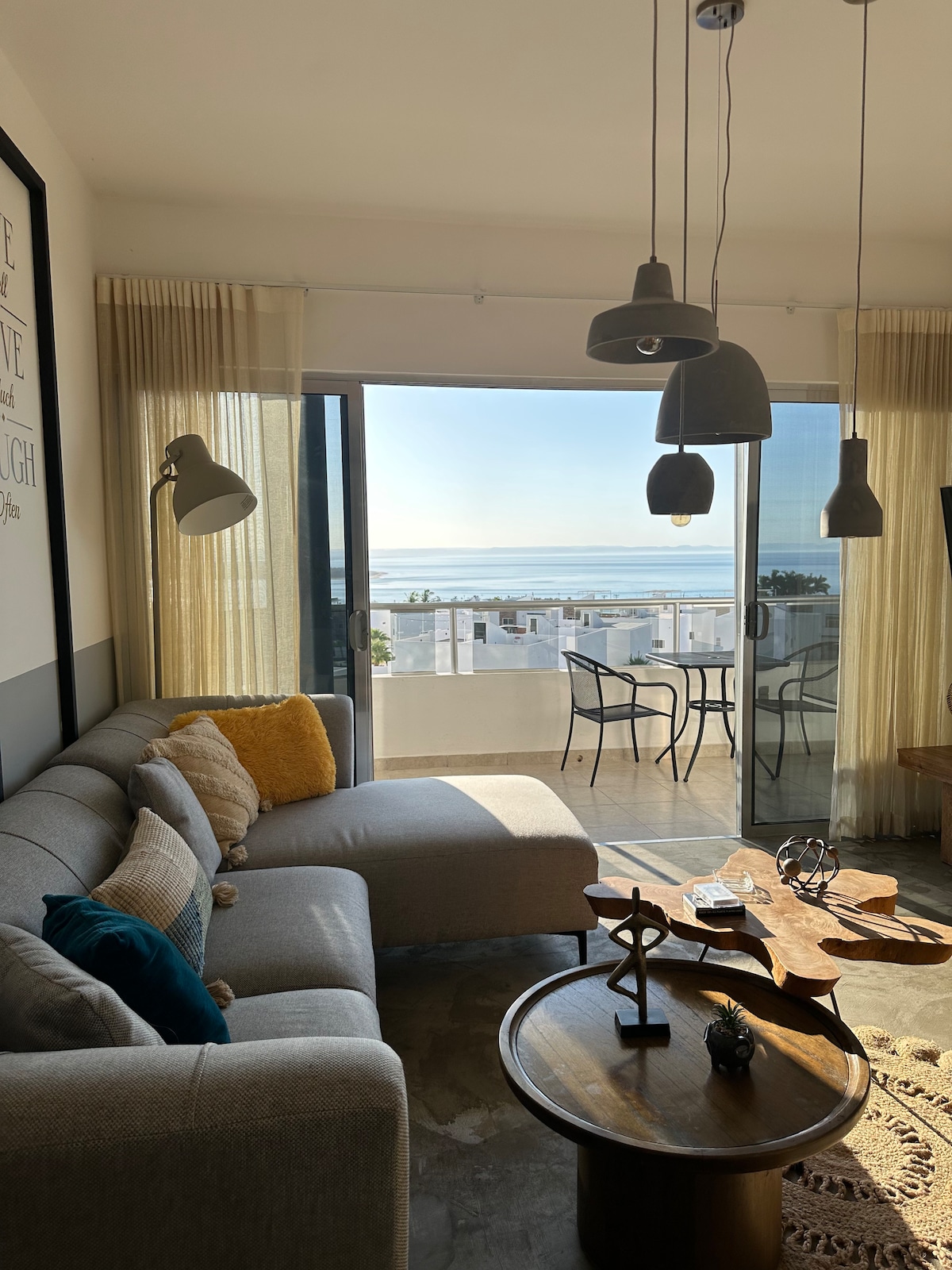 A cozy living area is displayed, featuring a gray sectional sofa with colorful cushions. A round coffee table sits nearby, complemented by decorative items. Large windows offer views of the ocean, and a small balcony table with chairs is visible outside.