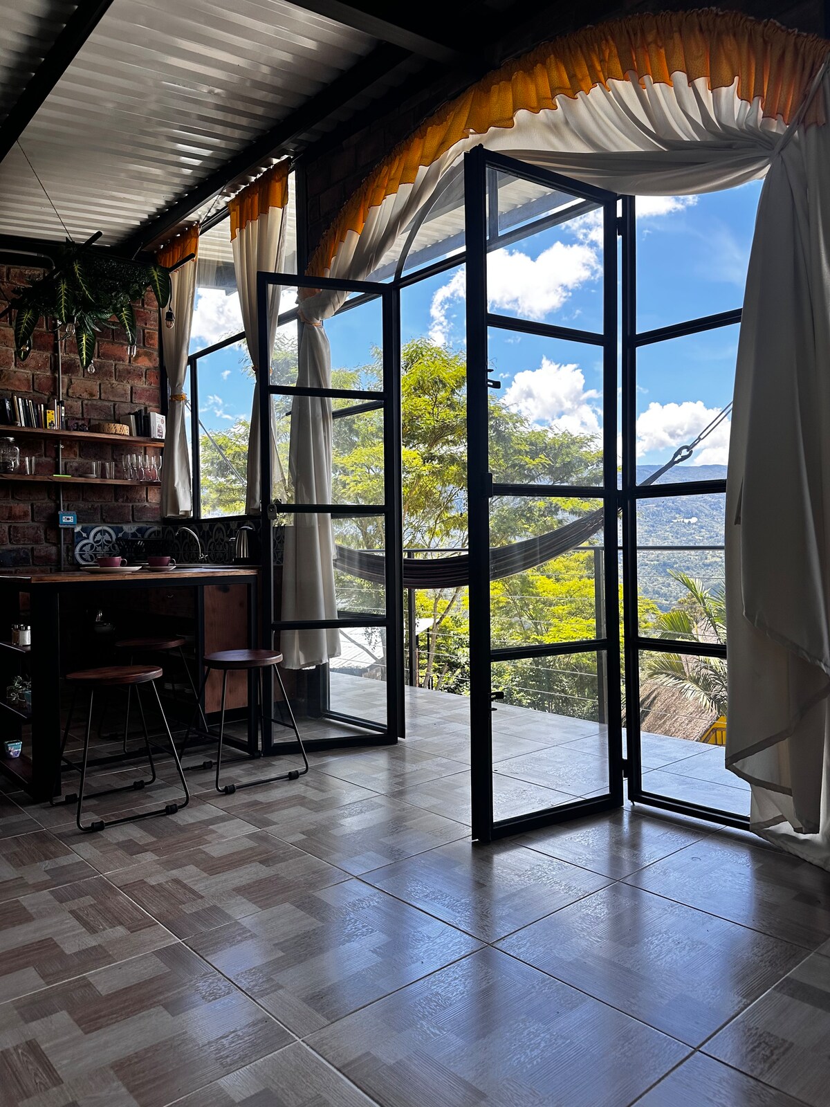 A welcoming dining area features a wooden table surrounded by high stools. Large glass doors open to reveal views of the surrounding greenery and a bright sky, allowing natural light to fill the space. Soft drapes frame the entrance for an airy ambiance.
