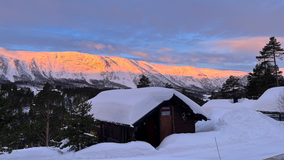 A snow-covered cabin is set against a backdrop of majestic mountains, illuminated by the soft hues of dawn. The landscape features gentle slopes and evergreen trees, while the sky displays a gradient of colors, enhancing the serene winter scene.