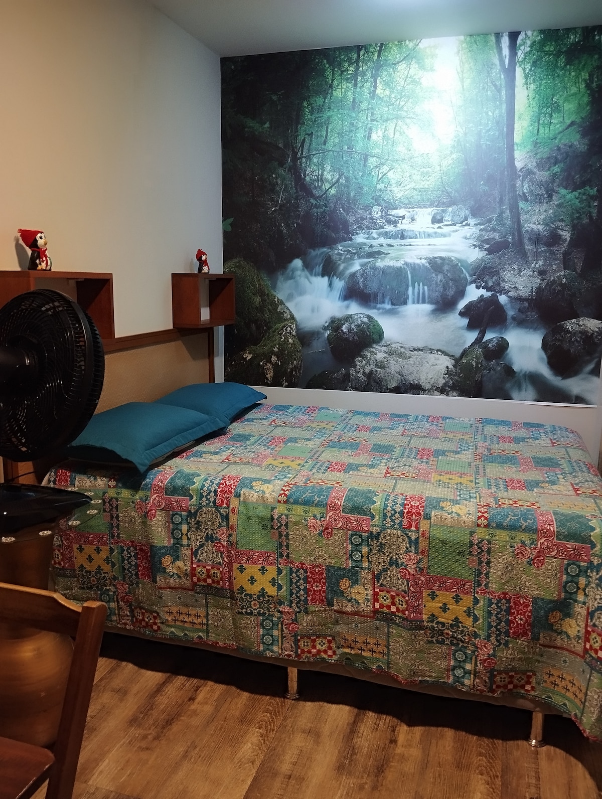 A comfortable sleeping area is complemented by a vibrant patchwork quilt draped over a spacious bed. A forest mural depicting a serene stream enhances the ambiance. A fan is positioned nearby, and simple wooden shelves hold decorative items, creating a cozy and inviting space.