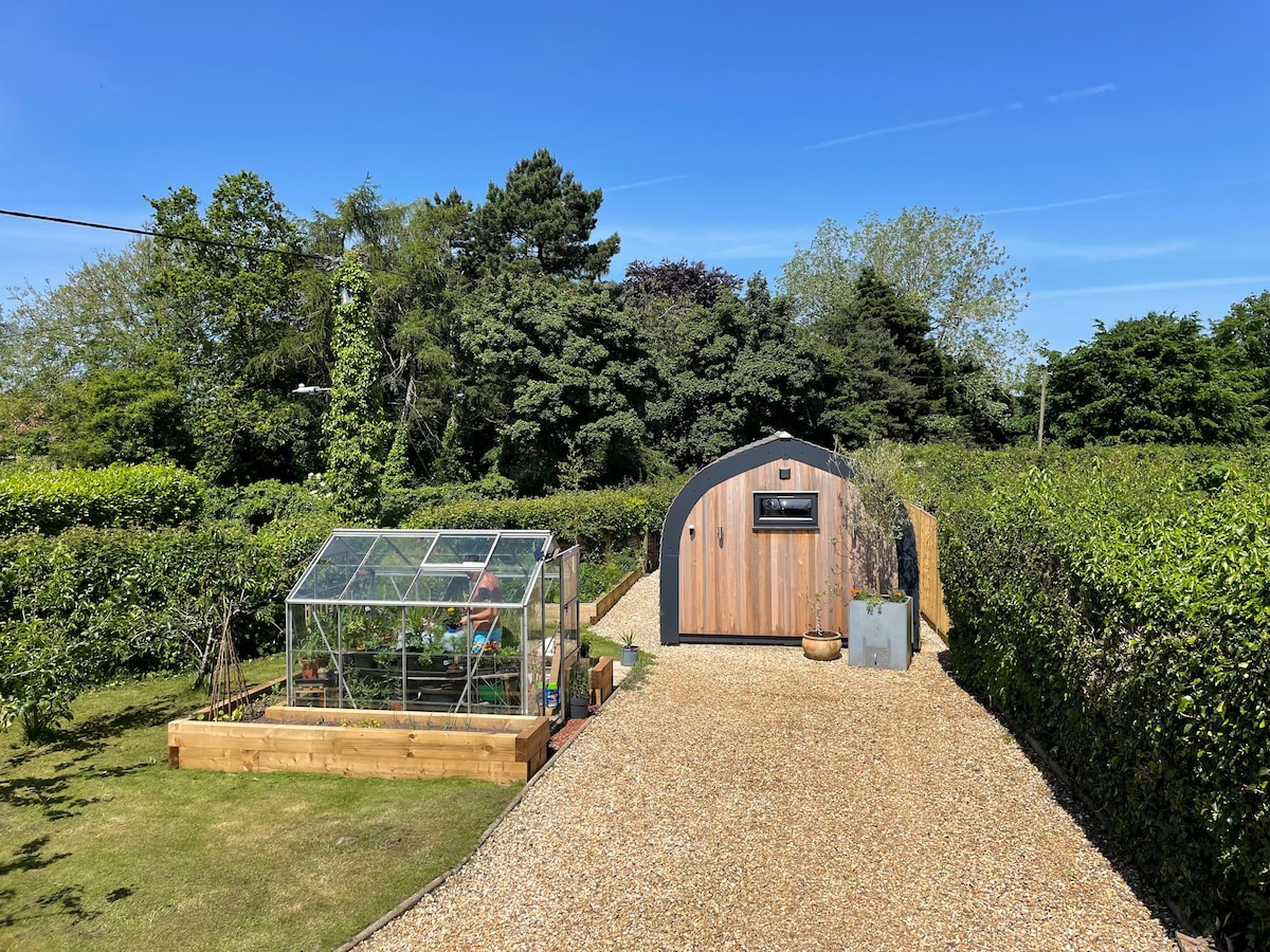 A gravel pathway leads to a wooden lodge with a curved roof, surrounded by lush greenery. A greenhouse is visible on the left, providing a contrasting structure within the landscape. Clear blue skies can be seen above, enhancing the natural setting.