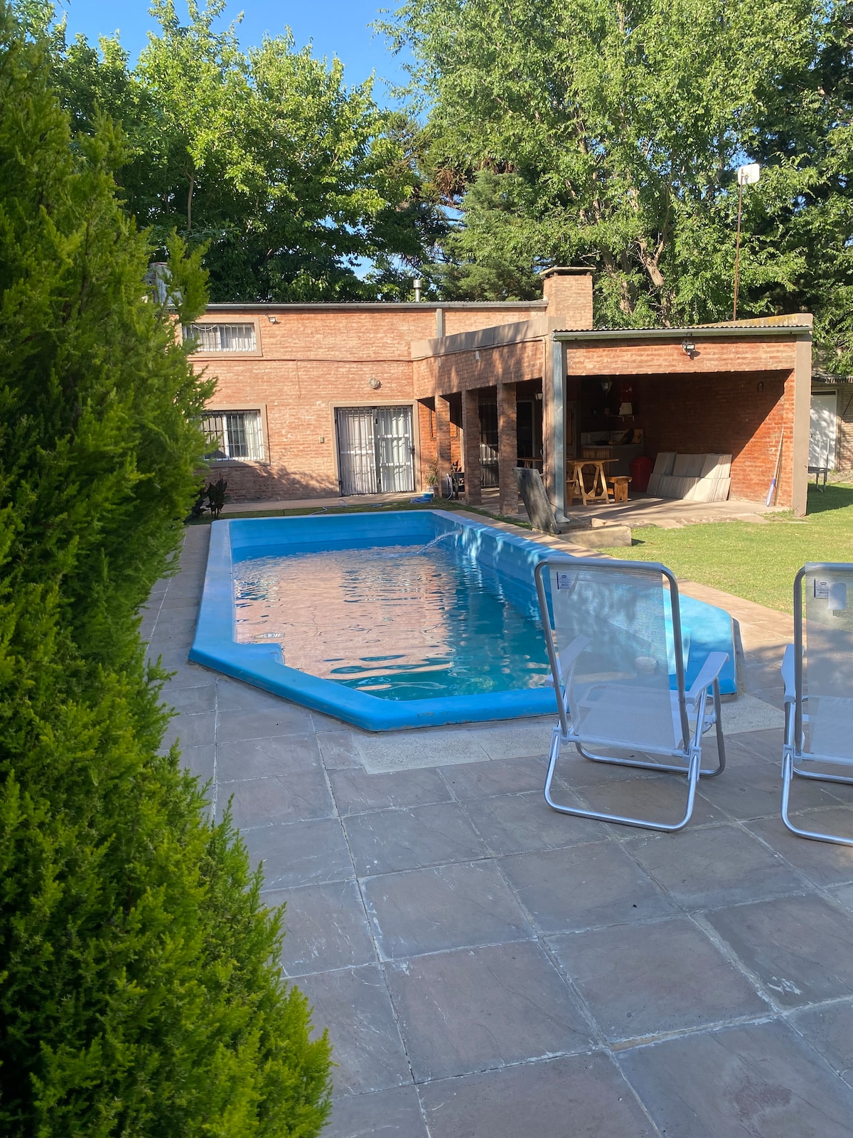 A serene outdoor space features a private rectangular pool surrounded by a paved terrace. Two light blue lounge chairs are positioned near the water. A grassy area extends away from the pool, with a brick house and shaded patio visible in the background.