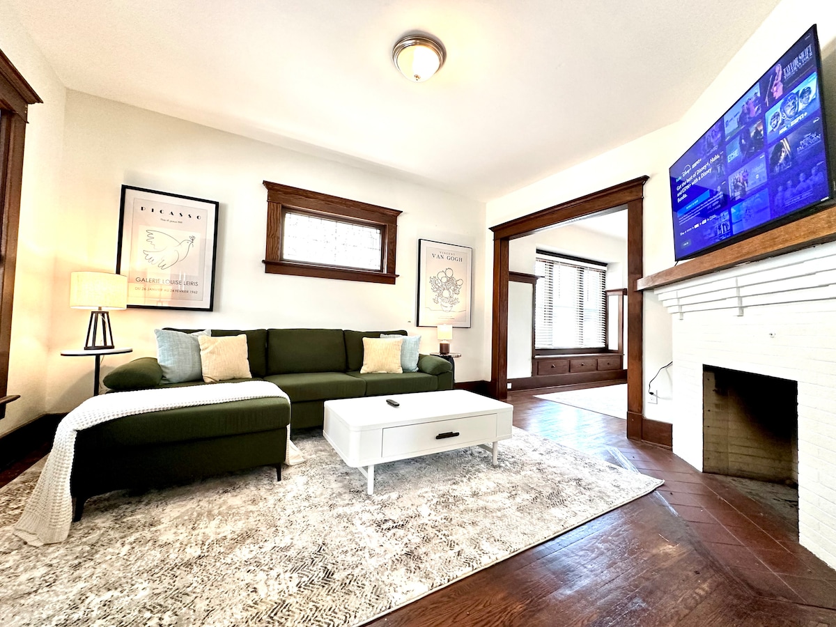 A comfortable living area is centered around a green sofa facing a modern coffee table. A television on the wall displays content. Light wood accents frame the space, complemented by large windows allowing natural light to fill the room.