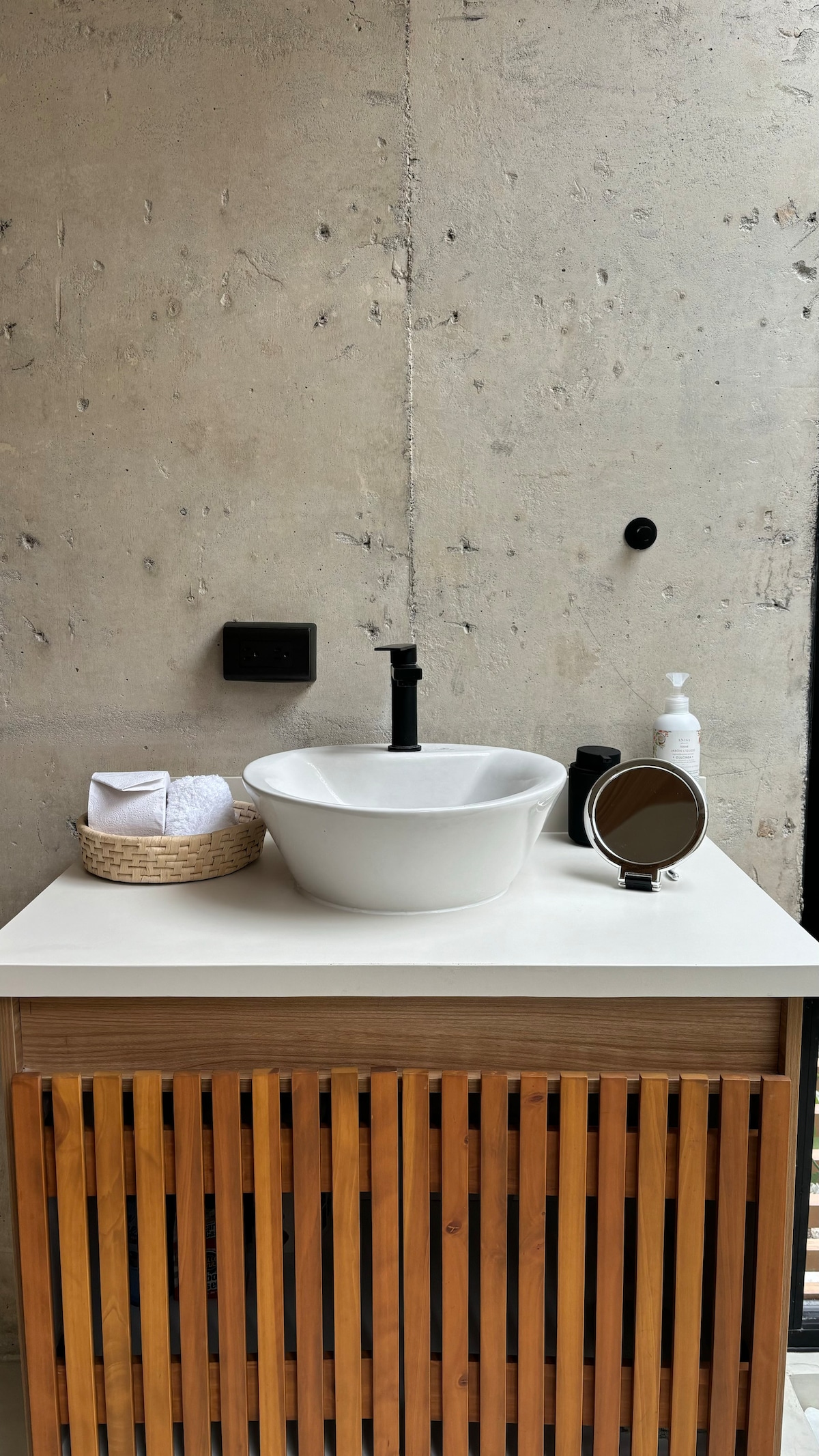 A clean and minimalist bathroom area features a round white vessel sink atop a light-colored counter. A woven basket contains neatly rolled towels, while a small mirror and an elegant soap dispenser are visible. The textured concrete wall enhances the modern aesthetic.