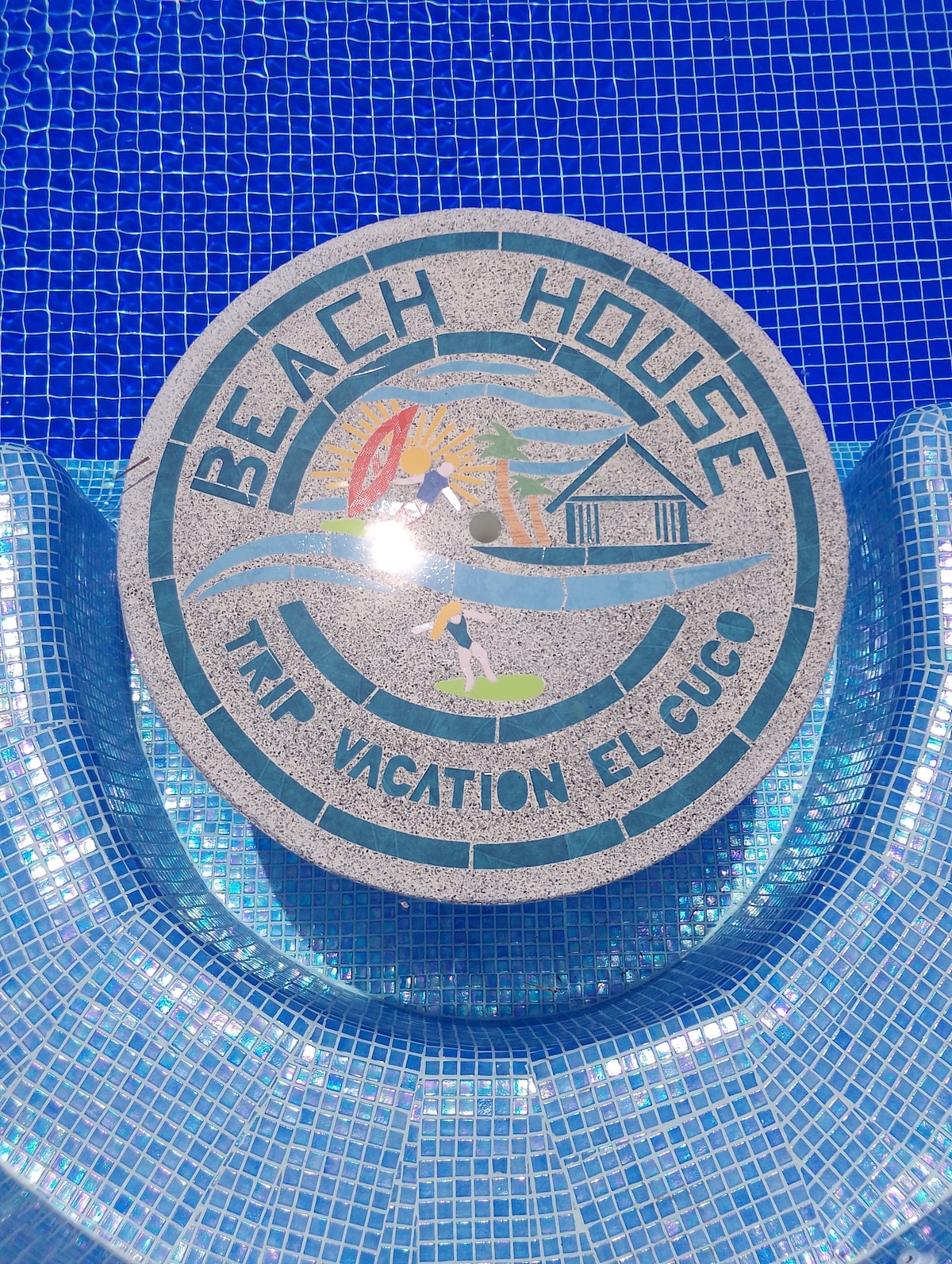 A circular stone emblem is displayed on the pool's edge, featuring the text 'BEACH HOUSE' along with a design showcasing water sports and a house. The logo is set against a backdrop of shimmering blue tiles, reflecting sunlight from the pool's water.