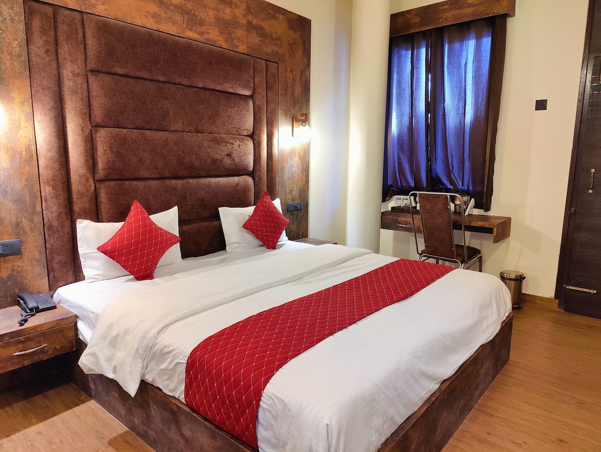 A spacious sleeping area features a large upholstered headboard in rich brown tones. The bed is adorned with two red accent pillows and a white bedspread. A work desk with a chair sits beside a window, which is dressed with dark curtains.