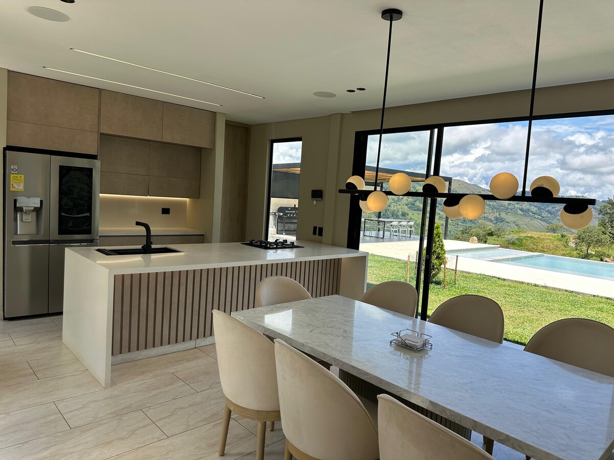 A modern kitchen and dining area features a spacious white countertop and light-colored cabinetry. A dining table surrounded by upholstered chairs is positioned near large windows, showcasing scenic views of the outdoor landscape and a pool. Soft lighting is provided by a contemporary pendant fixture.