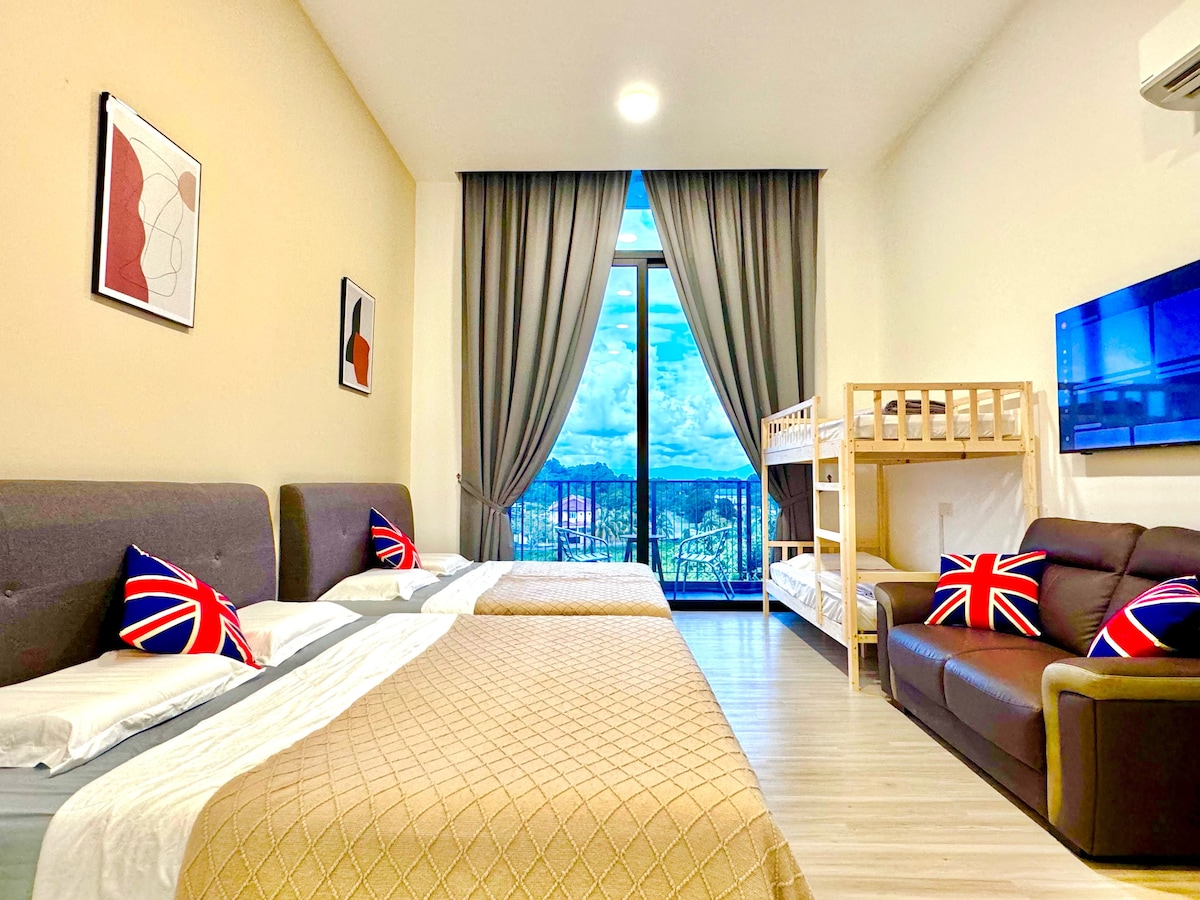 A spacious studio apartment features two large beds and a bunk bed, accented by patterned cushions. A sectional sofa is positioned in one corner. Large windows with sheer curtains provide natural light and views of the surrounding landscape, while a flat-screen TV is mounted on the wall.