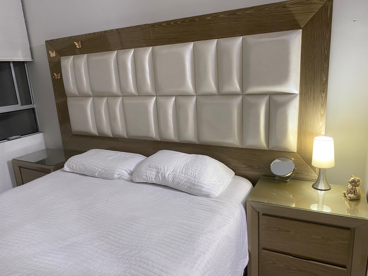 A spacious bed is positioned against a stylish headboard featuring a soft, padded finish. Flanking each side of the bed, bedside tables support decorative lamps and additional items. Soft, ambient lighting creates a calm setting, enhancing the room's modern design.