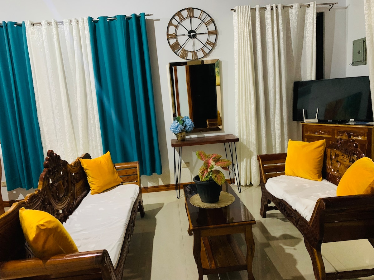 A cozy living area features two wooden seating arrangements with white cushions and yellow accent pillows. A wooden coffee table sits in the center, with a decorative plant placed on it. Tall curtains in teal and light fabric frame a large mirror, while a television stands nearby.