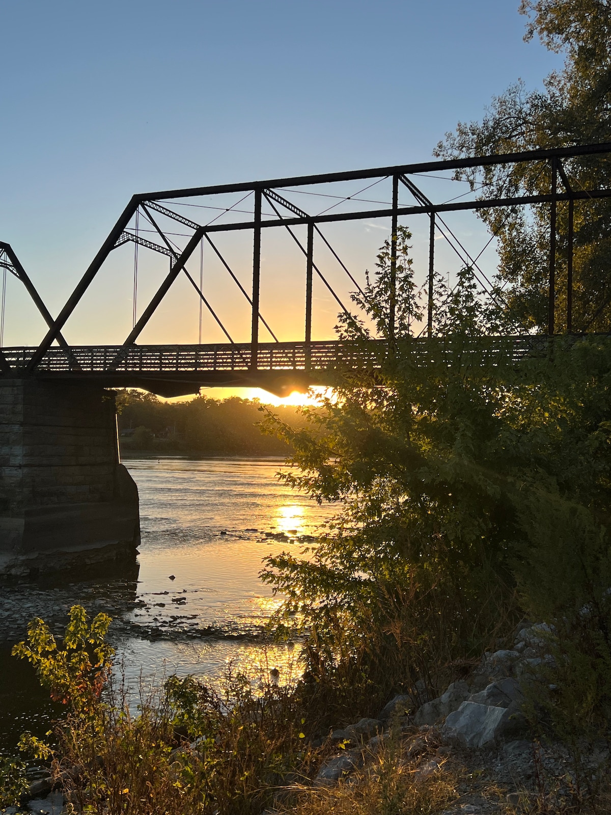 A historic metal-truss bridge spans a river, silhouetted against a vibrant sunset. Sunlight reflects on the water, creating a warm glow. Lush greenery and rocky banks frame the scene, enhancing the natural beauty of the landscape.