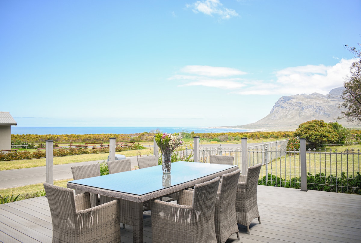 An outdoor dining area is featured with a large table surrounded by comfortable chairs. A vase of flowers decorates the center. In the background, expansive views of the ocean and rolling hills are visible under a clear blue sky.