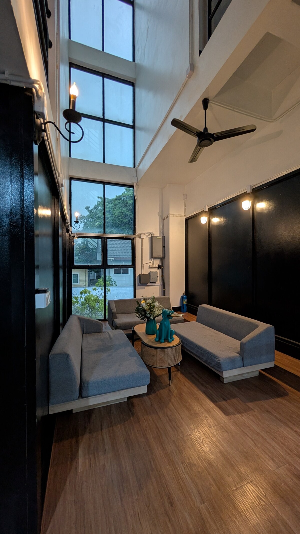 A bright lounge area features high ceilings and large windows, allowing natural light to fill the space. Two gray sofas are arranged around a round coffee table, complemented by decorative plants. An overhead fan and ambient lighting contribute to a comfortable atmosphere.