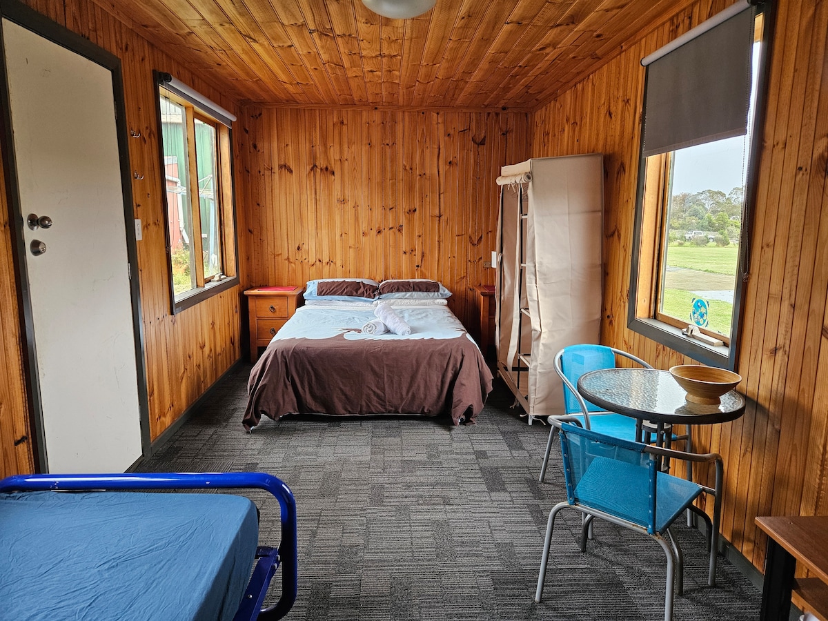A wooden interior cabin features a double bed covered with a brown blanket and a single bed with a blue frame. Sunlight enters through two windows, illuminating a small table with blue chairs and a chest of drawers.