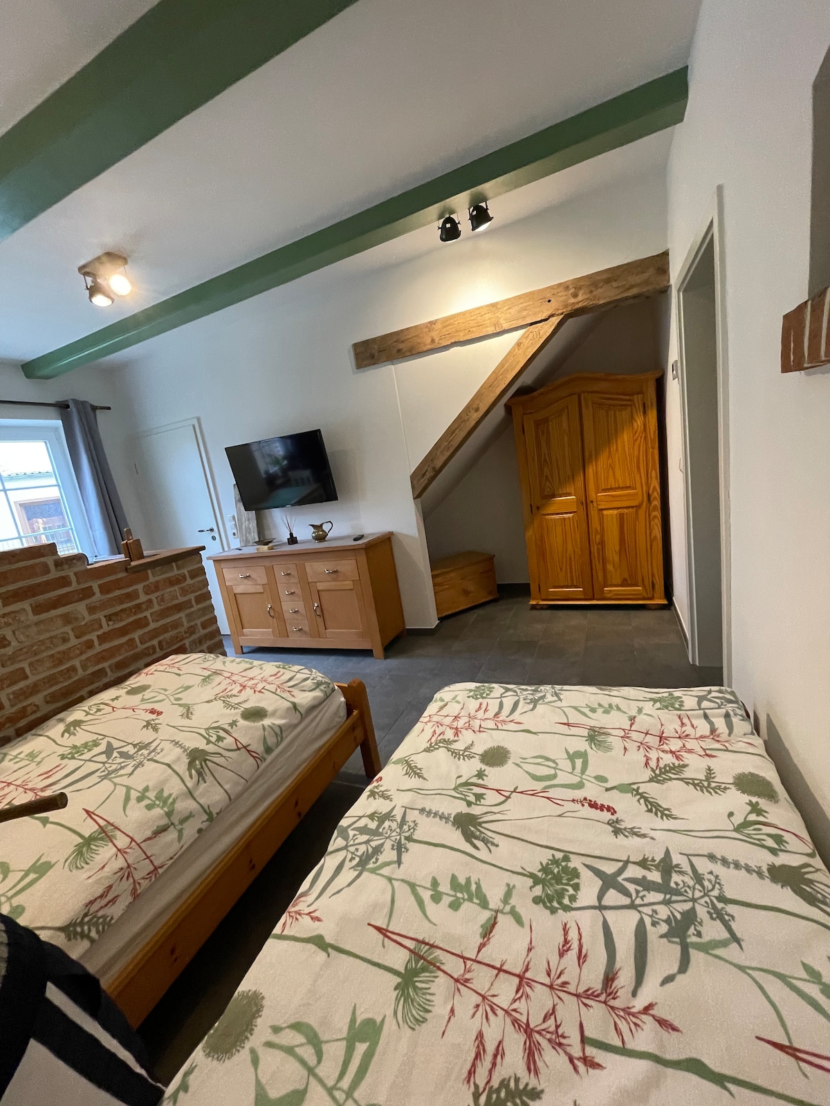 A serene sleeping area is presented with two individual beds featuring patterned bedding. Natural light enters through a window, illuminating wooden beams on the ceiling. A wooden wardrobe and a media console with a television are situated against a neutral wall, enhancing the room's functionality.