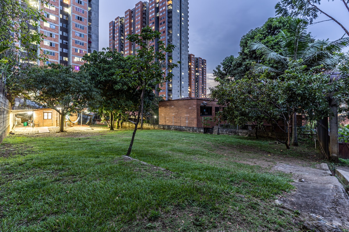 A spacious outdoor area is presented, featuring well-maintained green grass and a variety of trees. In the background, a rustic brick building is visible, and modern high-rise buildings flank the scene, emphasizing the blend of nature and urban living.