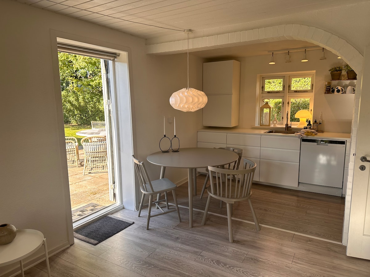 A dining area features a round table surrounded by three chairs, illuminated by a modern pendant light. A sliding door provides access to a garden view, while a well-equipped kitchen with white cabinetry and a dishwasher is visible in the background.