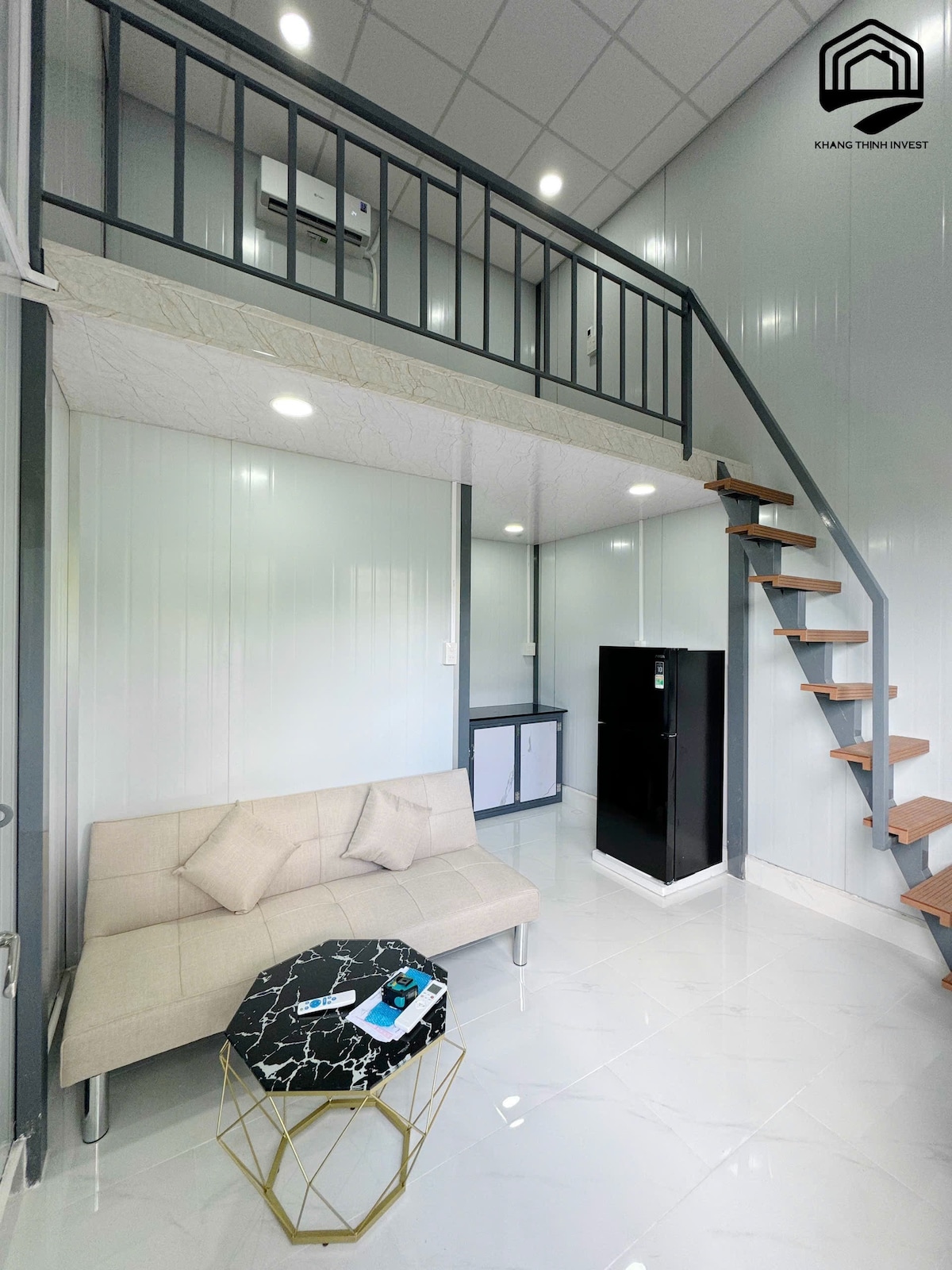 An interior view of a cozy mini house showcases a light-colored sofa facing an elegant coffee table. A staircase leads to an upper level. The kitchenette features a black refrigerator, while bright overhead lights enhance the modern ambiance of the room.