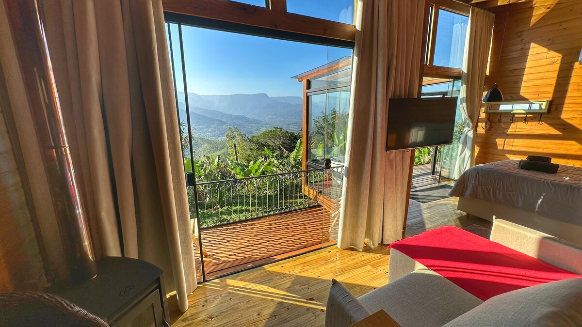 A spacious room features large glass doors that open to a stunning view of the mountains. Natural light floods the area, highlighting wooden flooring and warm wood accents. A comfortable seating area and a cozy bed are visible, creating an inviting atmosphere for relaxation.