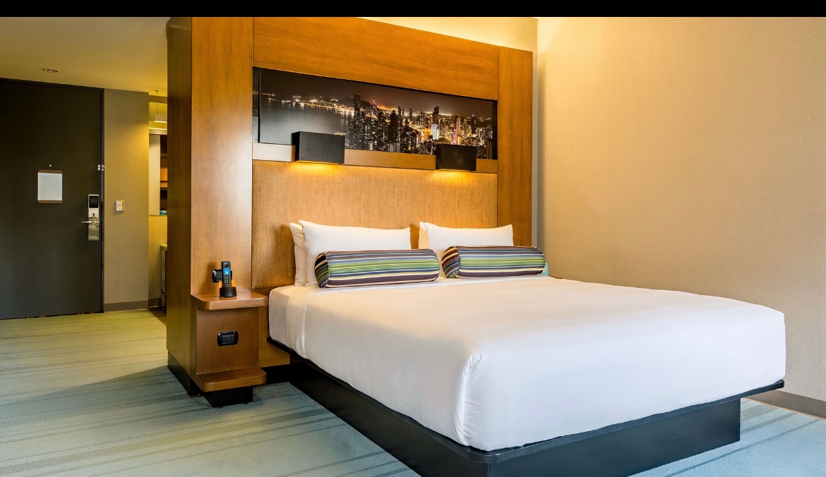 A spacious room features a king bed with soft white linens and colorful striped accent pillows. The headboard is constructed from light wood, complementing the contemporary design. A view of the city skyline is visible in the artwork above the bed, enhancing the room's ambiance.