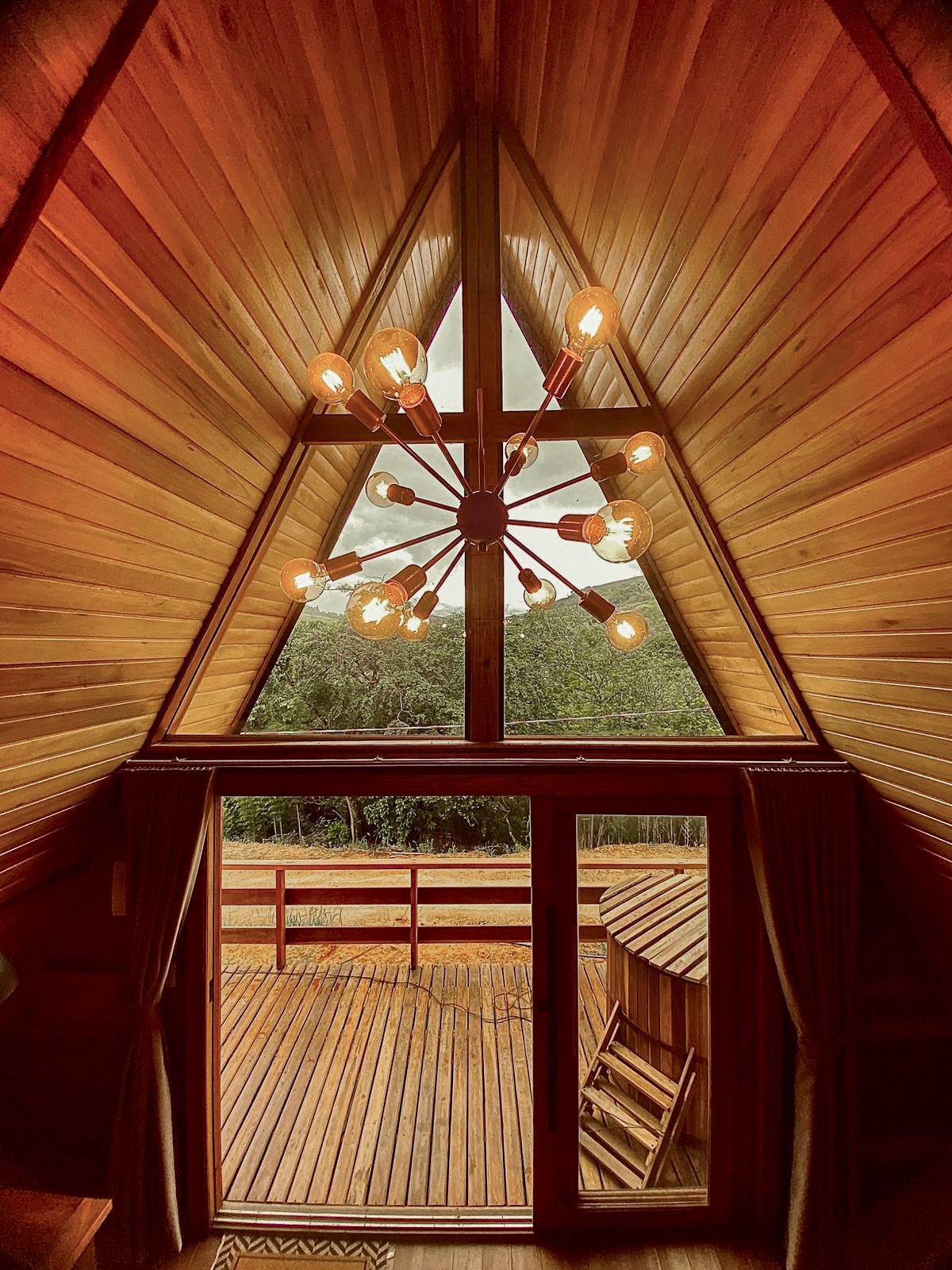 A triangular wood-paneled structure is showcased, featuring a striking chandelier with multiple bulbs. Large glass doors open to a wooden deck, providing a view of the outdoors. Natural light filters through the windows, enhancing the warm tones of the interior space.