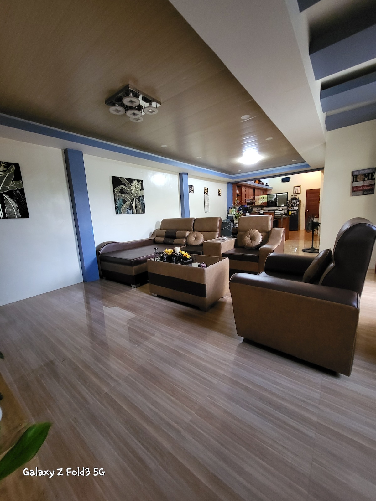A spacious living area showcases comfortable seating arrangements, including a large couch and two armchairs. A coffee table sits at the center, accentuated by decorative elements. Light filtered through the ceiling adds a warm glow to the inviting space, leading seamlessly to the kitchen area.