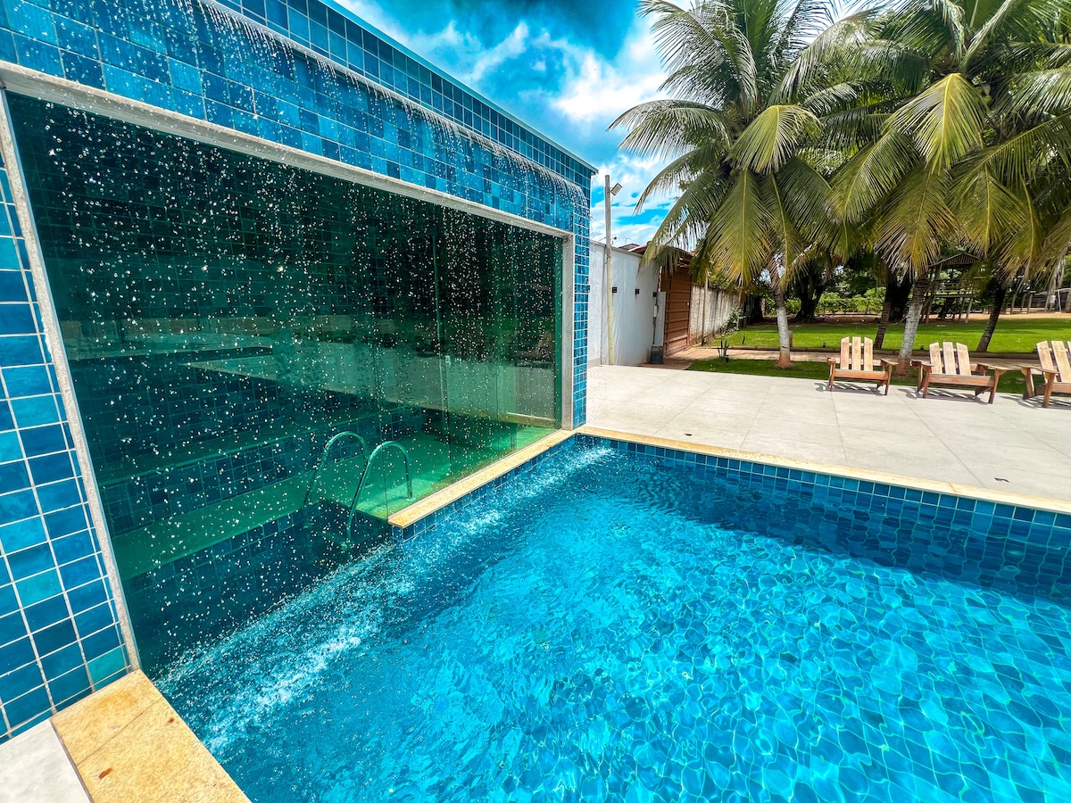 A vibrant swimming pool is featured with a cascading water wall, surrounded by blue tiles. Sunlight reflects off the clear water, accentuating the inviting atmosphere. Lounge chairs are positioned nearby, and palm trees provide shade in the background.