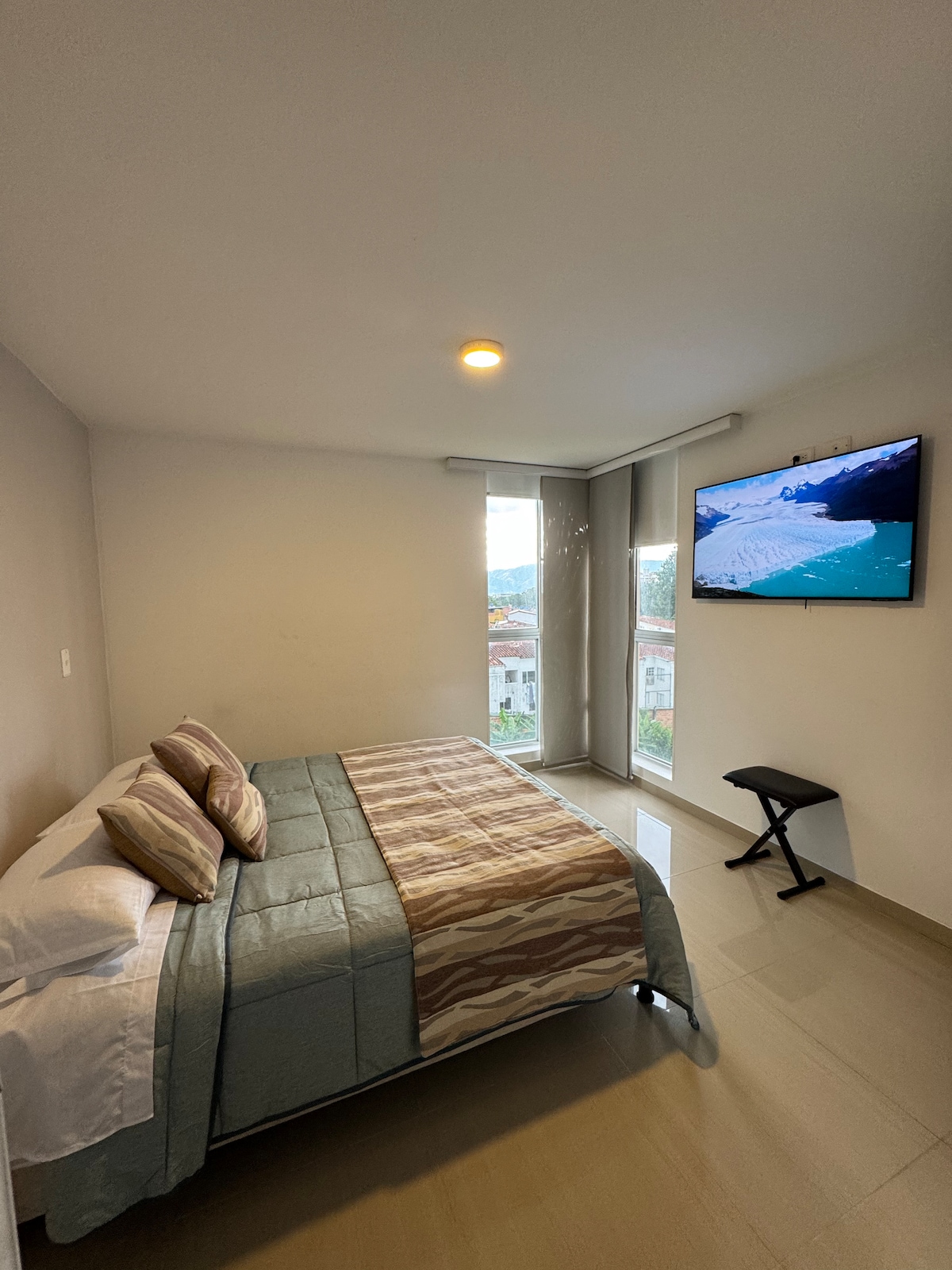An inviting sleeping area features a spacious bed with decorative pillows and a patterned coverlet. A flat-screen television is mounted on the wall, and two large windows allow natural light to enter, enhancing the room's brightness.
