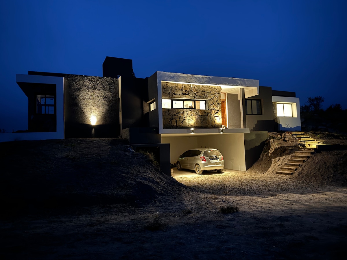 The modern exterior of the residence is illuminated at night, showcasing a blend of stone and contemporary architecture. A vehicle is parked in the spacious carport beneath the main entrance, with steps leading up to the front door. Warm lighting highlights the building's structure.