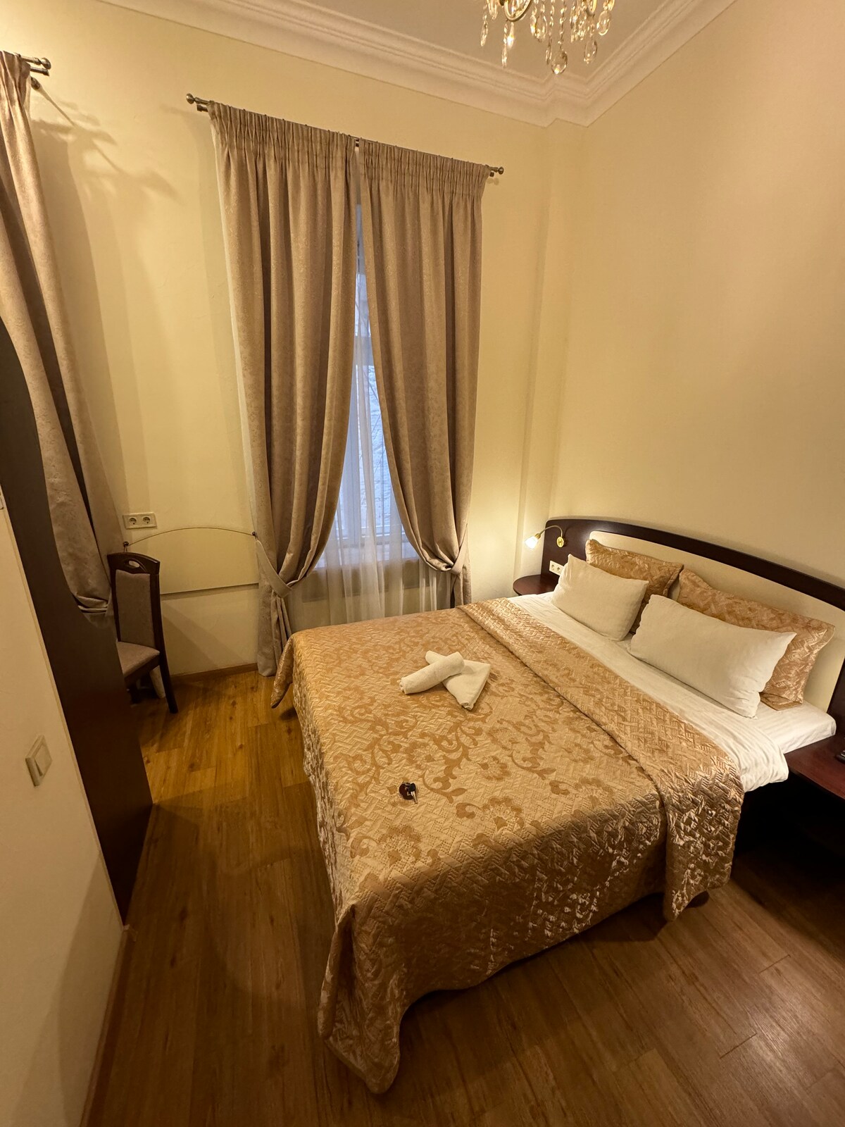 A cozy bedroom features soft beige tones, with a spacious bed adorned with decorative pillows and rolled towels. A pair of elegant curtains frame the window, allowing natural light to softly illuminate the room. A minimalist chair is positioned near the wall.