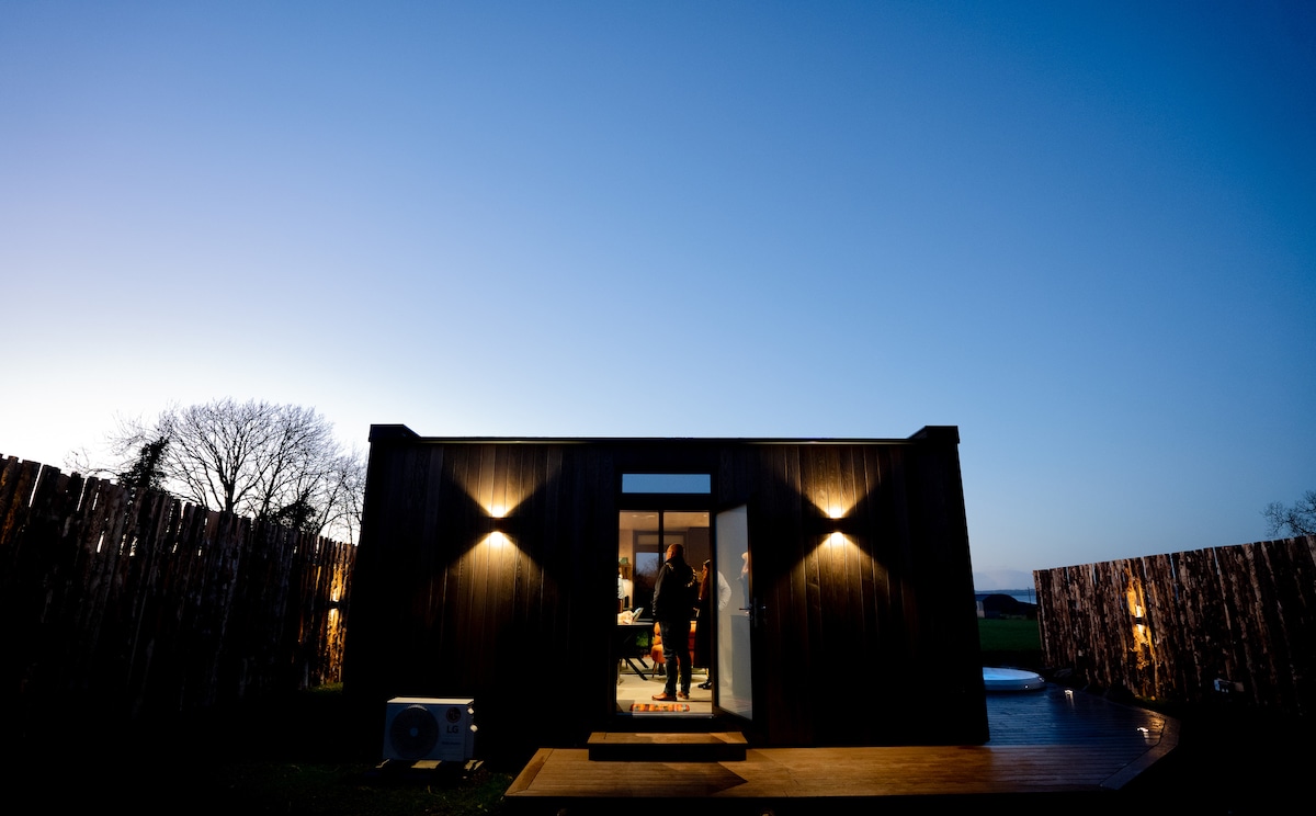 A modern, mirrored cabin stands against a twilight sky, featuring soft exterior lighting on either side of the entrance. The structure is surrounded by rustic wooden fencing, with a glimpse of the landscape beyond, creating a peaceful ambiance.
