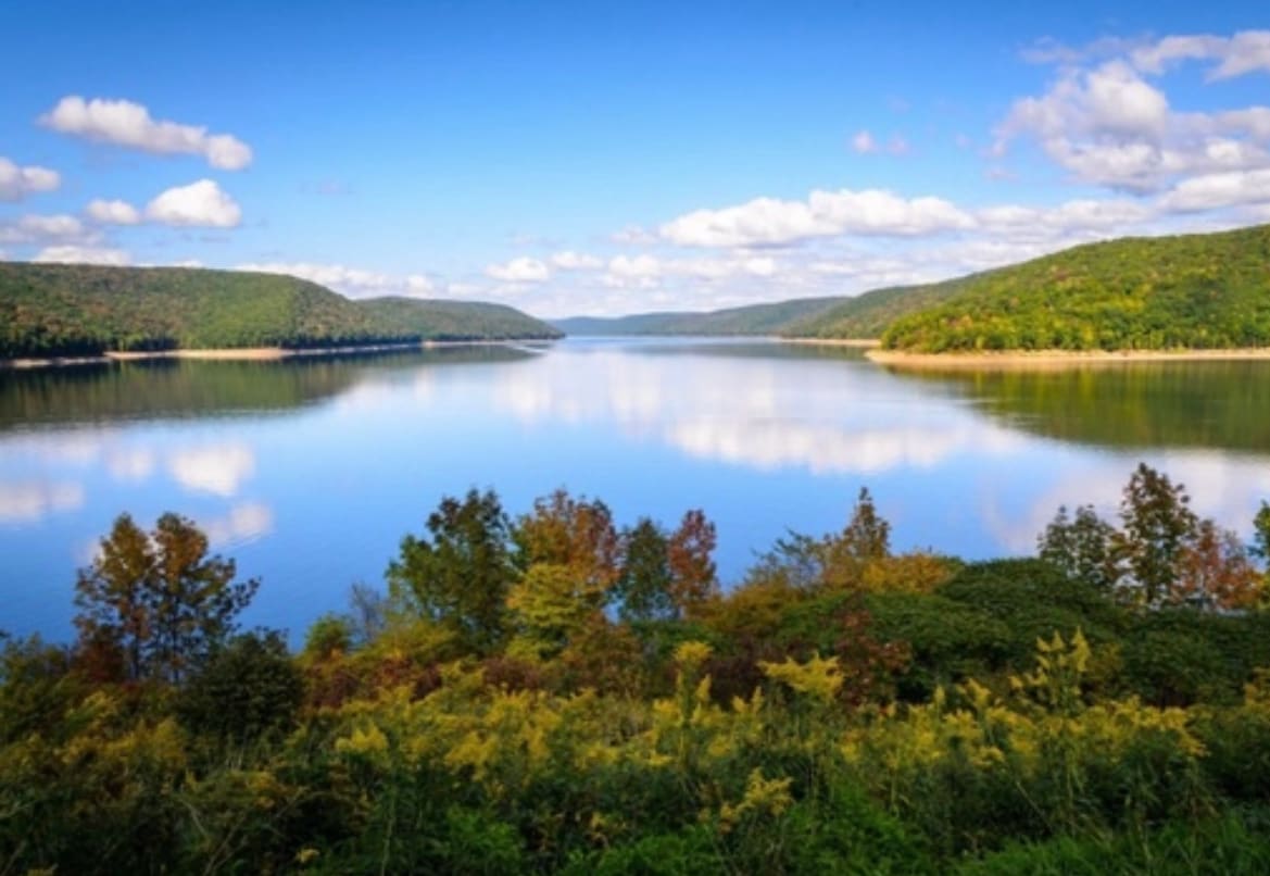 A serene lake view is captured, featuring calm waters reflecting the sky and surrounding green hills. Soft clouds drift overhead, while a variety of trees line the shore, showcasing autumn colors of orange and yellow, creating a peaceful natural setting.