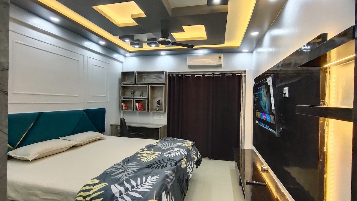 A modern bedroom features a spacious bed adorned with a patterned bedspread. Soft lighting highlights the crafted ceiling design. A dark curtain provides privacy, while a wall-mounted television and a desk area add functional elements to the space.