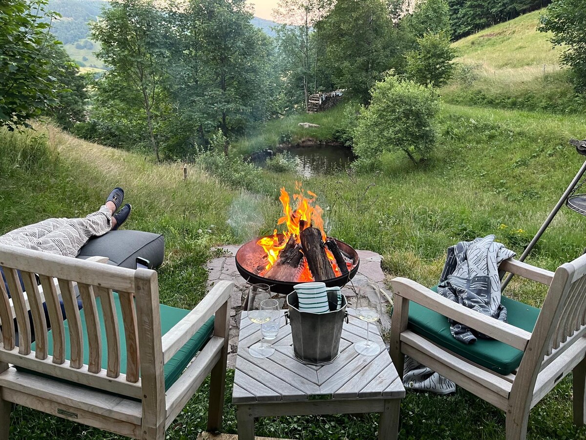 A fire pit is positioned at the center of a grassy area, surrounded by two wooden chairs with green cushions. A small table holds a bucket and plates, while a bubbling brook and trees create a natural backdrop. The serene atmosphere invites relaxation.