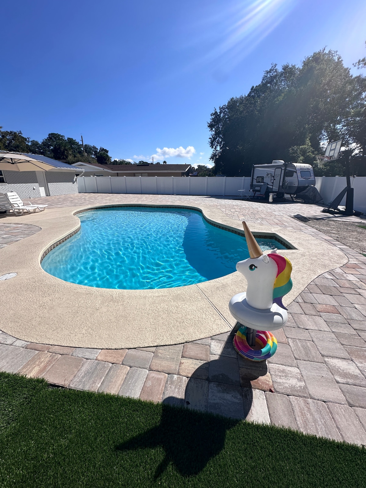 A private pool area displays a sparkling blue pool, surrounded by a sunbathing deck. A colorful unicorn float rests at the pool's edge, contrasting with the light-colored patio surface. White lounge chairs are positioned nearby, providing space for relaxation under a clear sky.