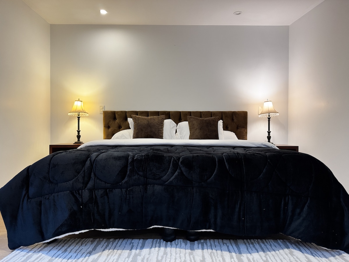 A spacious bed is framed by soft, neutral walls, featuring a plush headboard and decorative pillows. Two bedside lamps provide gentle lighting, enhancing the calming ambiance of the room. A textured blanket lays across the bed, while a light area rug adds warmth to the floor.
