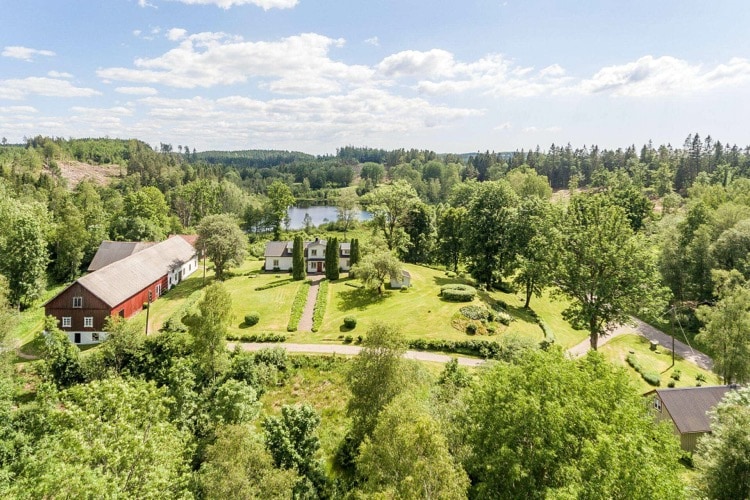 An aerial view captures a serene landscape featuring a lush green yard with scattered trees. A cluster of buildings, including a red barn and several houses, is visible, alongside neat pathways leading towards a calm lake framed by vibrant foliage.