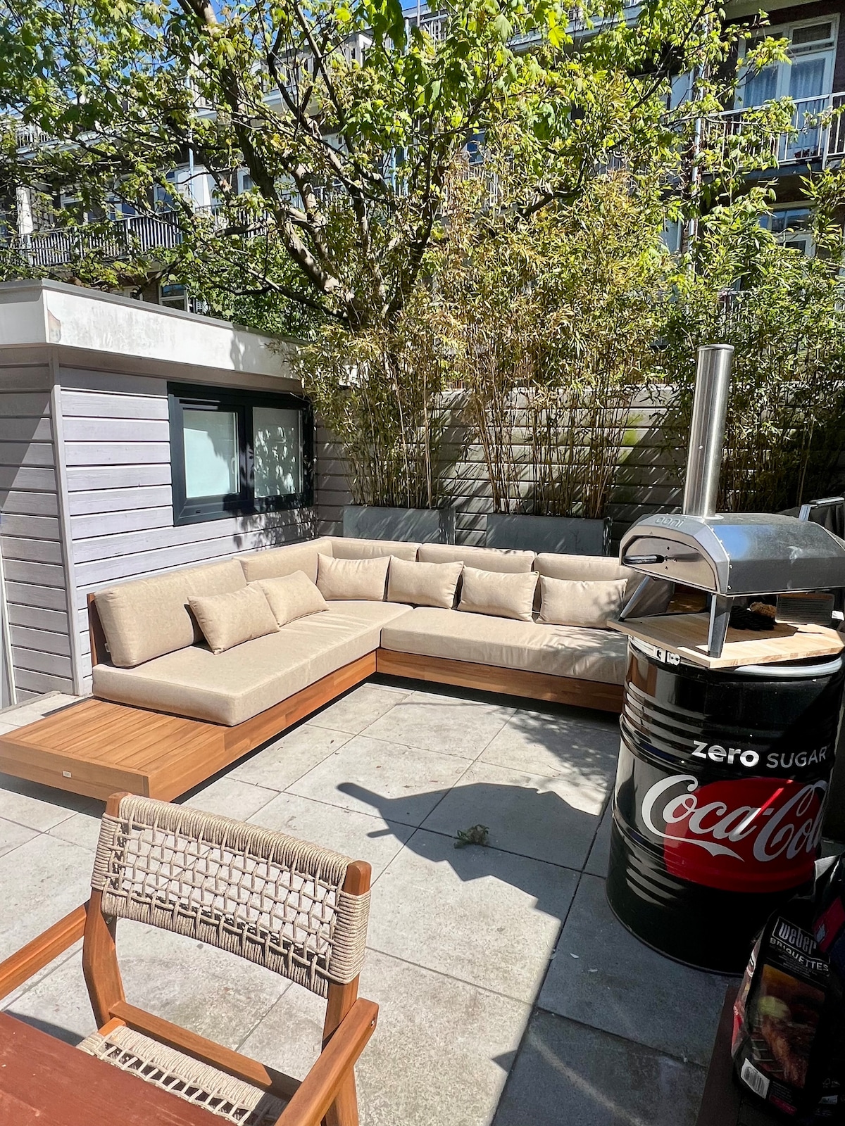 An outdoor seating area is showcased, featuring a spacious sectional couch with light-colored cushions arranged around a wooden deck. A metal grill stands beside a round table, complemented by a chair made of natural materials. Surrounding greenery adds a touch of privacy to the space.