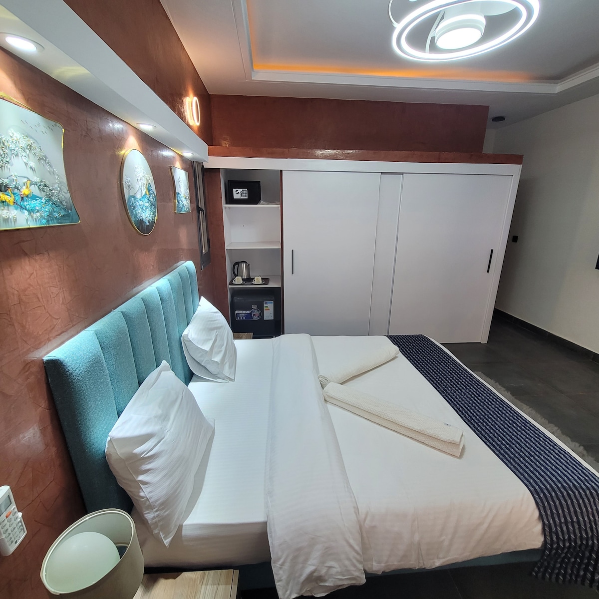 A well-lit sleeping area features a spacious bed dressed in white linens. Decorative artwork adorns the wall above the bed, and a sleek side table holds a lamp. A white wardrobe and a mini-fridge are visible in the background, contributing to a modern aesthetic.