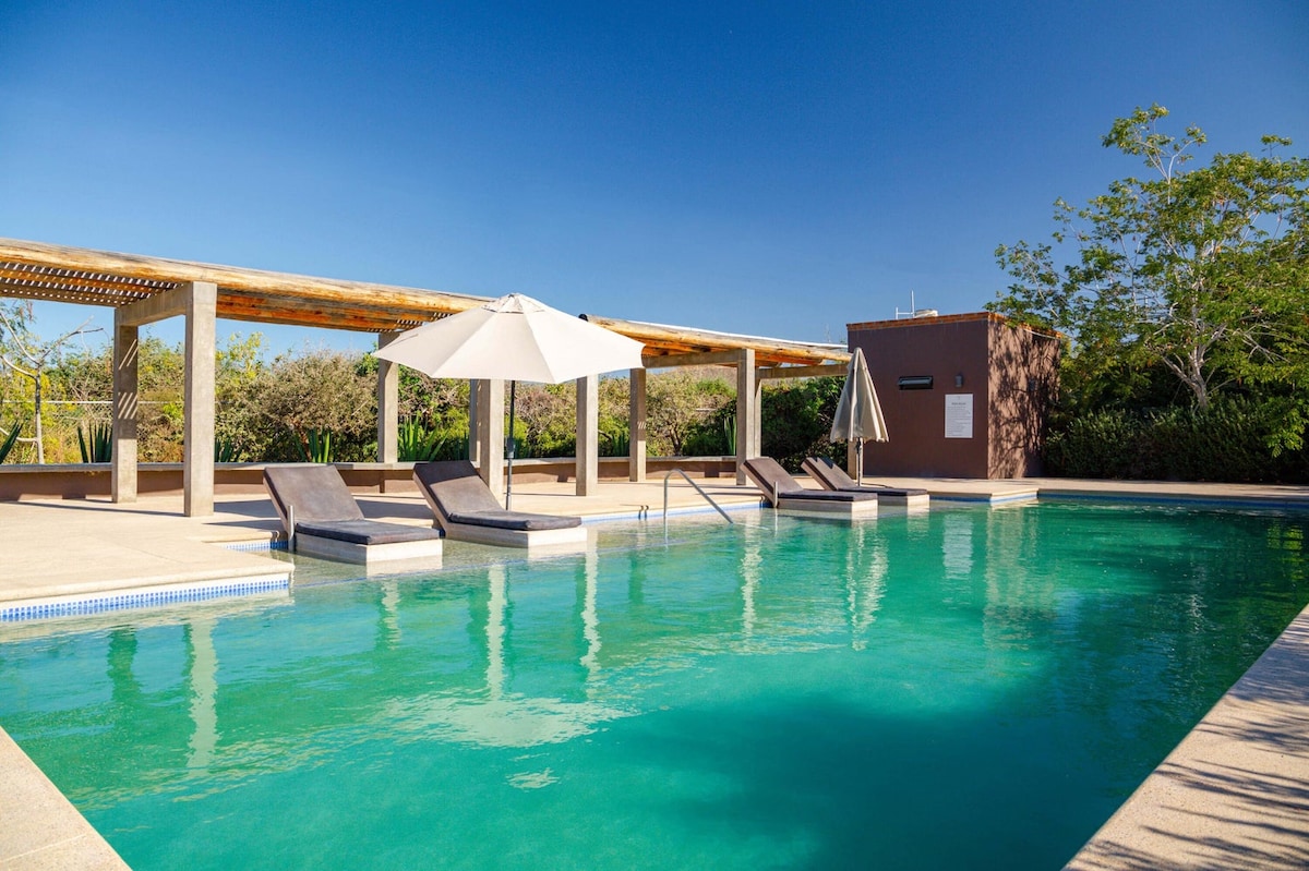 A heated saltwater pool is surrounded by a spacious deck featuring lounge chairs and umbrellas for shade. Lush greenery forms a natural backdrop, while a shaded area is provided for relaxation and gathering. The clear blue sky enhances the inviting atmosphere of this outdoor space.